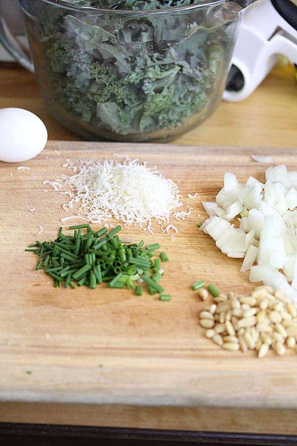 Ingredients for Kale Spanakopitas including an egg, parmesan, chopped white onion, pine nuts, and spring onions