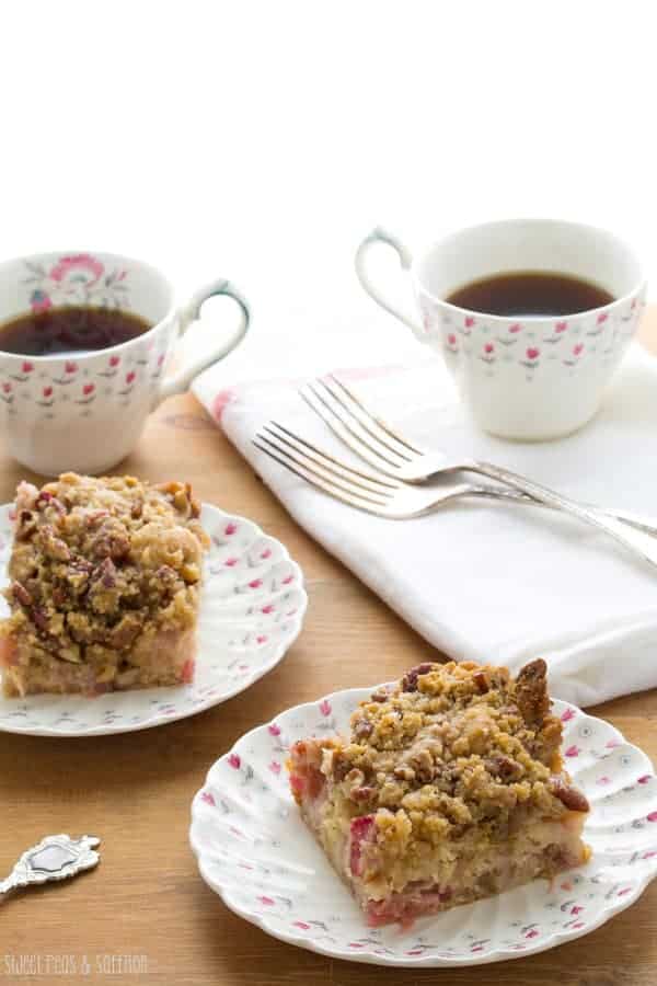 Two pieces of Rhubarb Coffee Cake with Pecan Streusel on plates with cups of coffee behind them