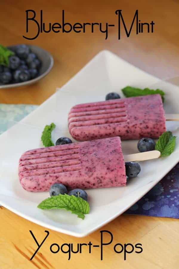 Two Blueberry Mint Yogurt Popsicles on a white plate with fresh blueberries and fresh mint as garnish