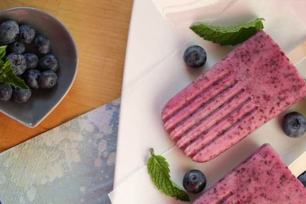 Overhead shot of Blueberry Mint Yogurt Popsicles on a white plate with bowl of fresh blueberries next to it