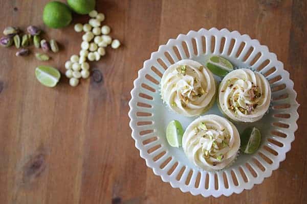 Three Key Lime Cupcakes on a cake stand with white chocolate chips, pistachios, and limes on the side