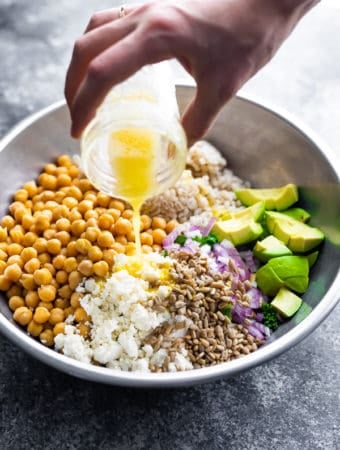 a hand pouring honey lemon vinaigrette onto a kale barley salad with feta and avocado