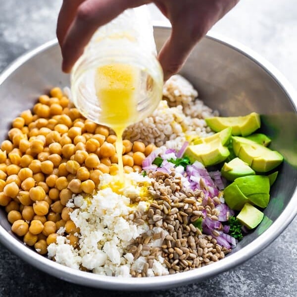 a hand pouring honey lemon vinaigrette onto a kale barley salad with feta and avocado