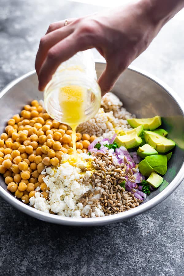 a hand pouring honey lemon vinaigrette onto a kale barley salad with feta and avocado