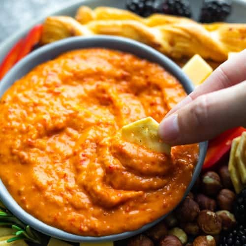 hand dipping a cracker into roasted red pepper dip in blue bowl