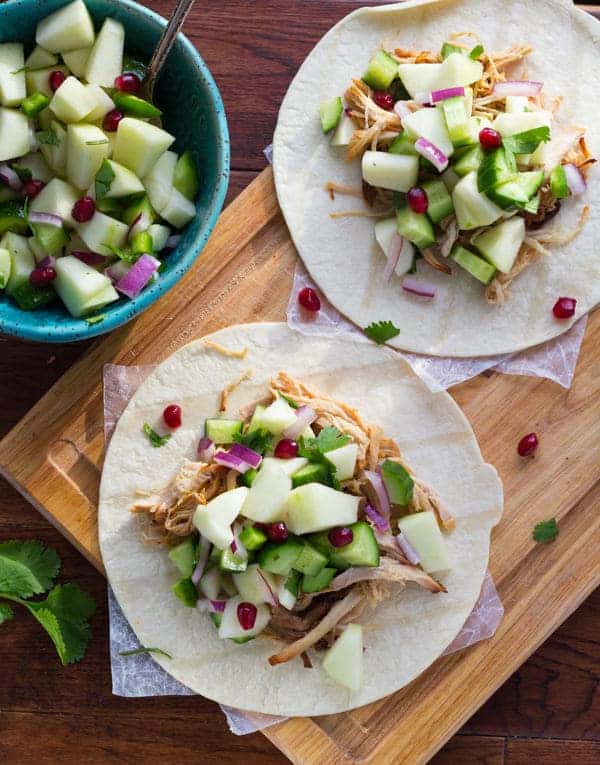 overhead shot of 2 tortillas topped with apple cider carnitas and fresh apple salsa on wooden cutting board and bowl of apple salsa to the side