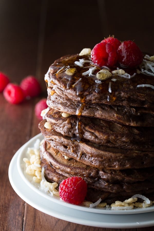 stack of chocolate rice krispie pancakes on plate with raspberries, shredded coconut on top and syrup drizzling down