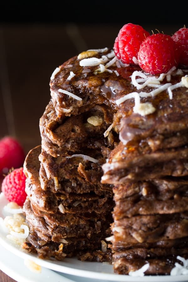 stack of chocolate rice krispie pancakes on plate with raspberries, shredded coconut on top and syrup drizzling down with slice taken out