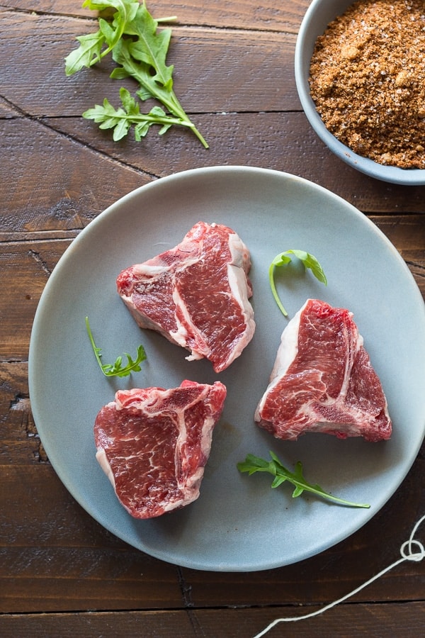 three uncooked lamb chops on a plate with a bowl of brown sugar chili rub nearby