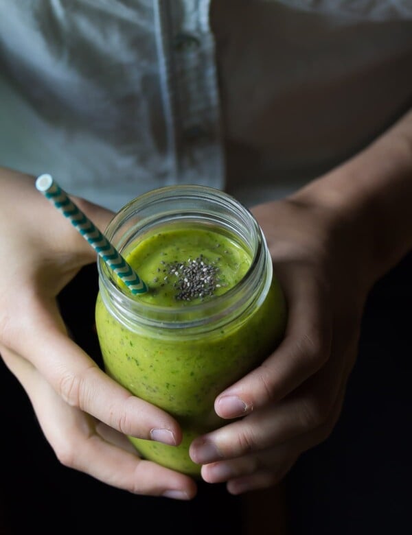 hands holding a green mango superfood smoothie in mason jar with blue straw
