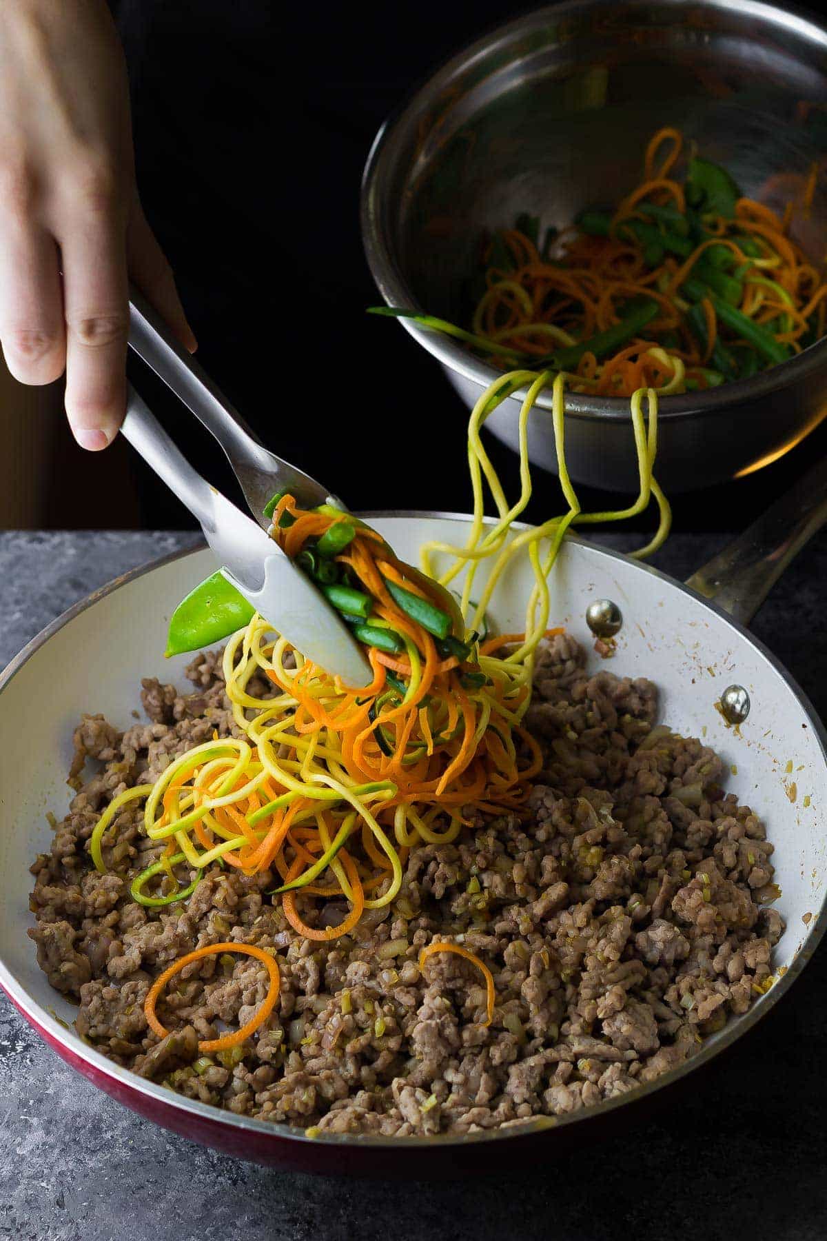 Stir fry vegetables being added into a skillet with cooked ground pork