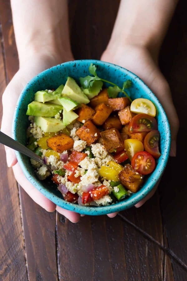 hands holding a blue bowl with tofu breakfast scramble with sweet potatoes and avocado