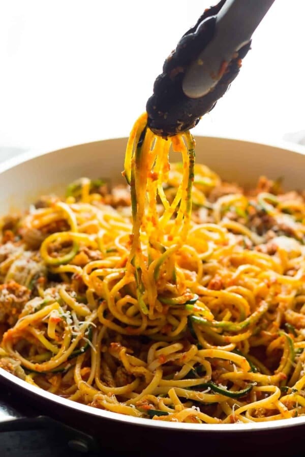 A bowl of ground turkey pasta in romesco sauce with tongs pulling some out of white bowl