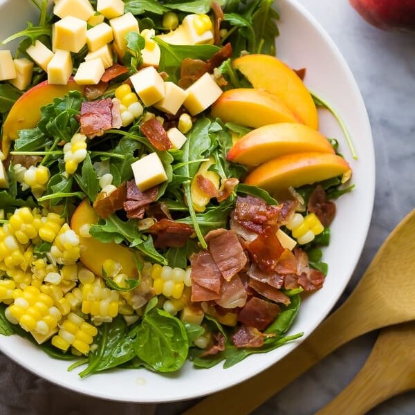 overhead shot of peach corn arugula salad in large white bowl with wooden tongs