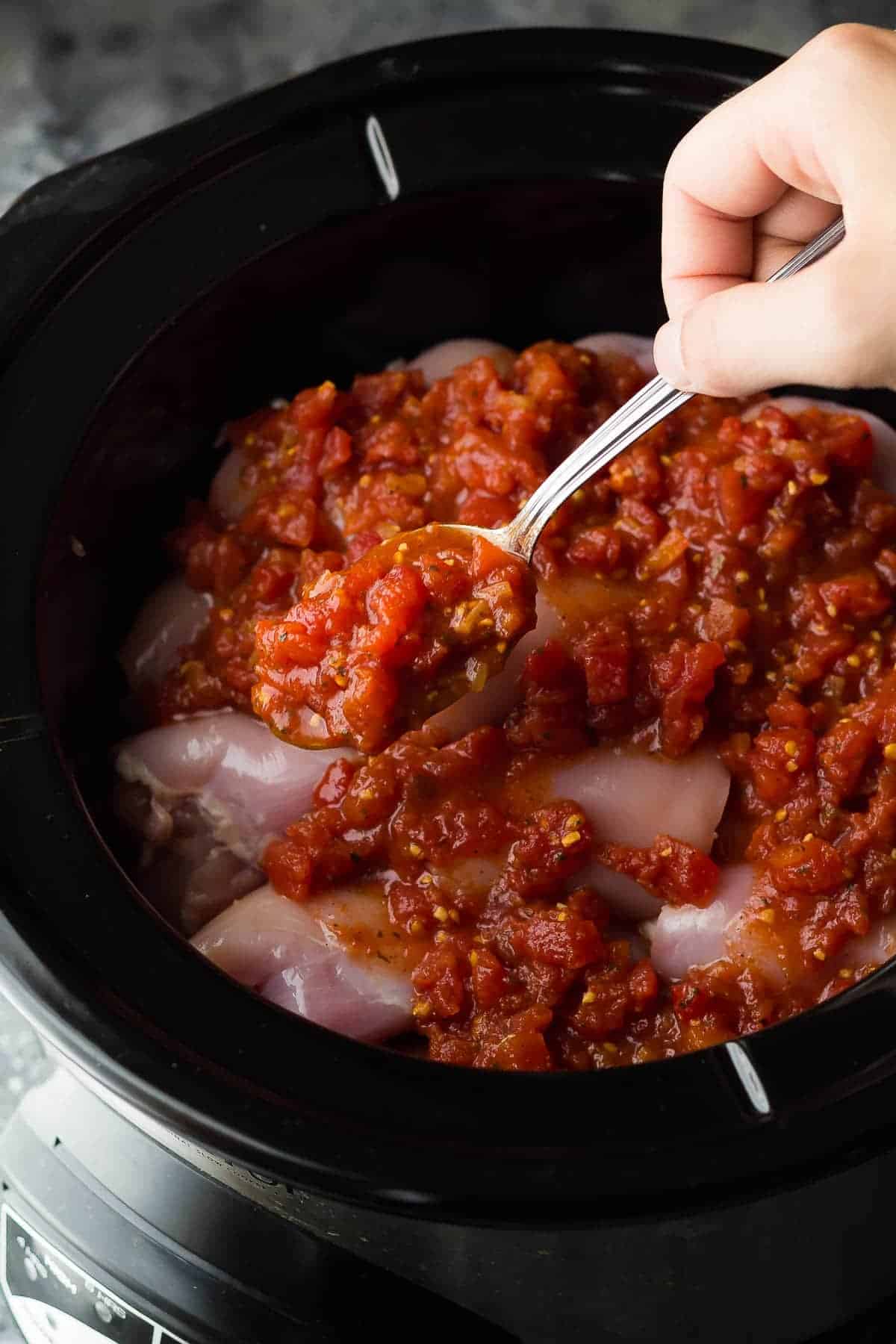 spooning diced tomatoes over chicken thighs in a slow cooker