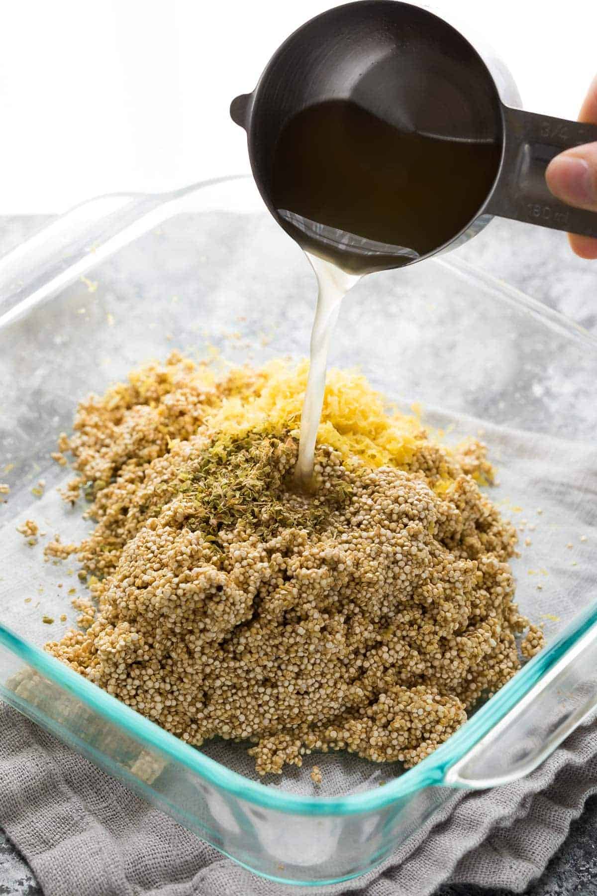 chicken stock being poured over quinoa, oregano, and lemon zest in glass casserole dish