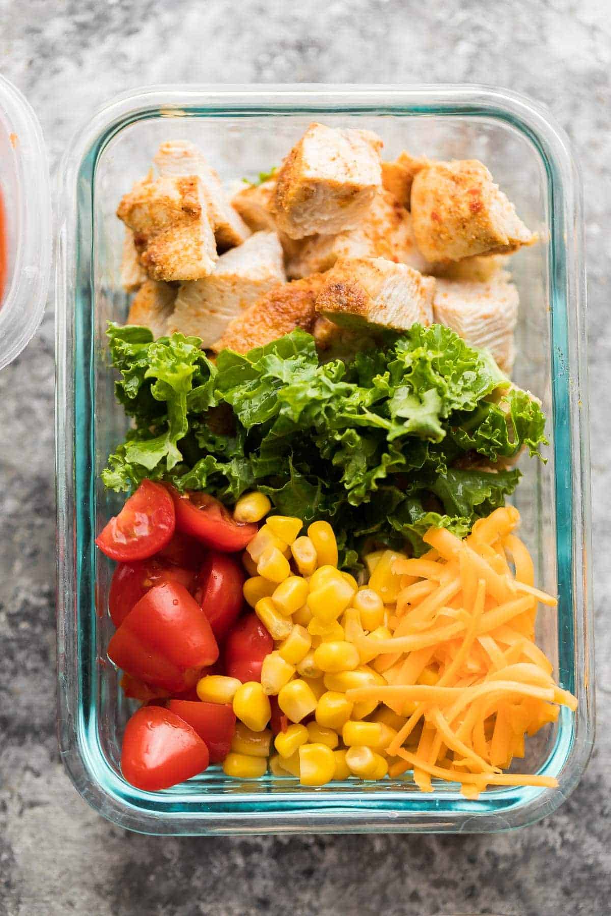 meal prep burrito bowl with chicken, tomatoes, corn, lettuce, and shredded cheese