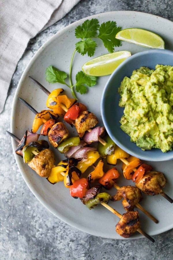 Overhead view of three fajita chicken skewers on gray plate with guacamole and lime wedges