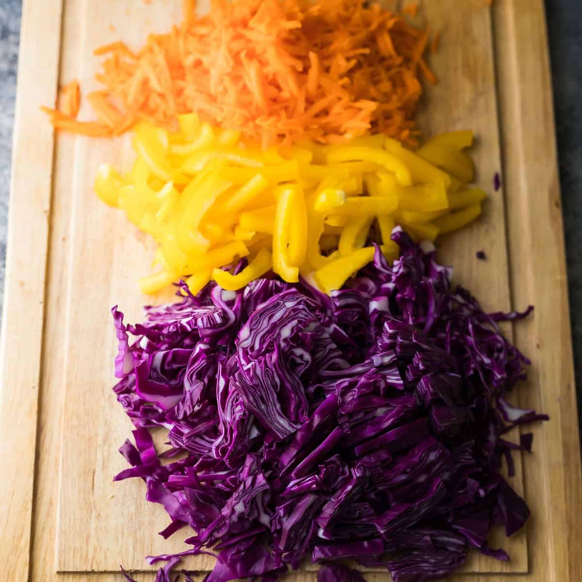 wood cutting board with shredded purple cabbage, sliced yellow pepper, and shredded carrots