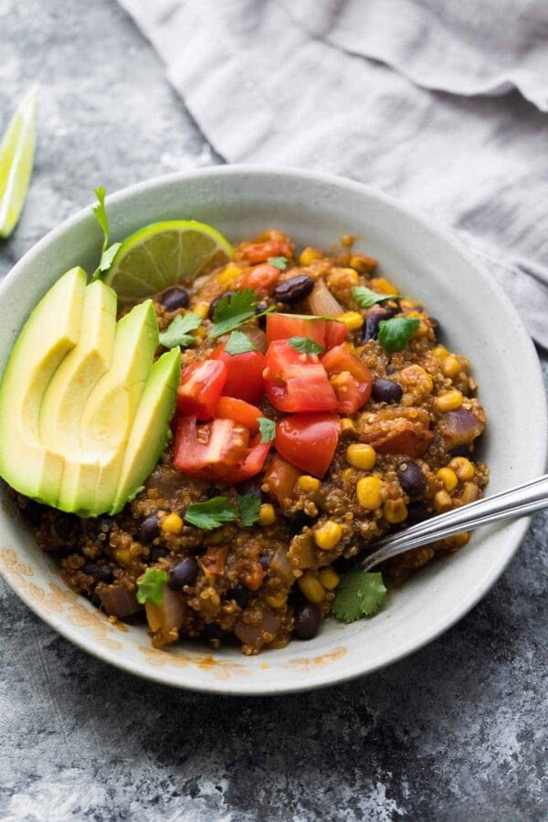 Quinoa Enchilada Casserole in a white bowl with avocado slices, tomatoes, lime wedges, and cilantro