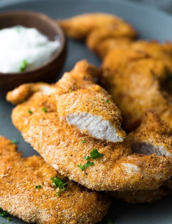 Crispy chicken tenders with a bite taken out of one and dipping sauce on a gray plate
