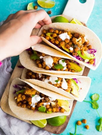 four air fryer cauliflower chickpea tacos lined up on blue background with hand grabbing one