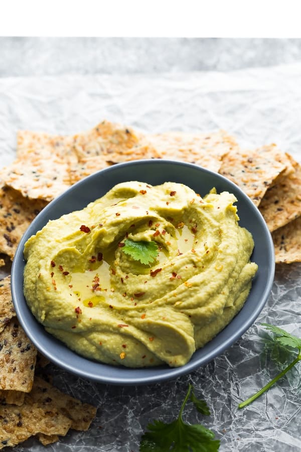 creamy avocado hummus recipe in blue bowl surrounded by crackers