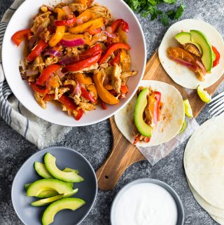 Overhead view of chicken fajitas in large white bowl with side of avocado slices, sour cream, tortillas, and wood cutting board