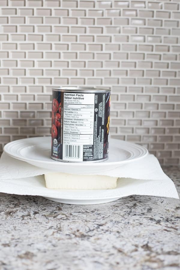 Side view of tofu on a white plate with a paper towel on it and a can sitting on top of the plate to drain tofu