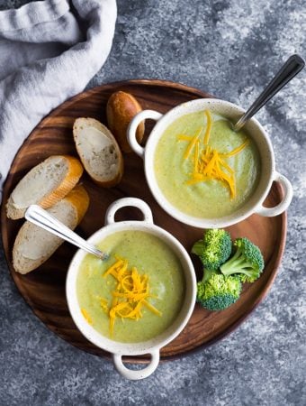 overhead shot of healthy broccoli cheese soup in two white bowls with fresh bread, cheese and broccoli on wood tray