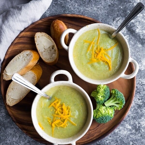 overhead shot of healthy broccoli cheese soup in two white bowls with fresh bread, cheese and broccoli on wood tray