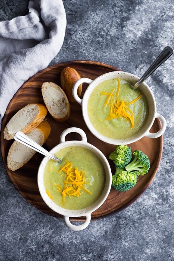 overhead shot of healthy broccoli cheese soup in two white bowls with fresh bread, cheese and broccoli on wood tray