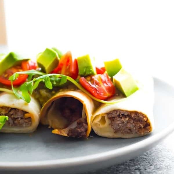 three baked beef taquitos on white plate with fresh avocados and tomatoes