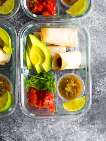 overhead shot of baked beef taquitos in glass meal prep container with fresh avocado, tomato, and lime