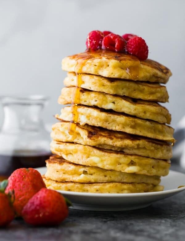 Side view of stack of cottage cheese pancakes on white plate with syrup and raspberries