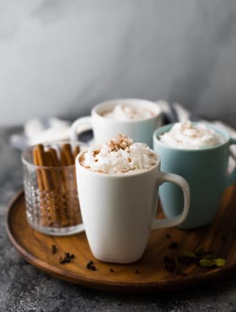 three cups of chai tea latte and a cup of cinnamon sticks on wood tray