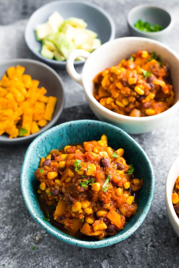 chili in blue bowl with bowls of avocado, cilantro, and corn chips in background