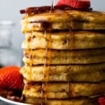 side view of a large stack of bacon pancakes on a plate with syrup being drizzled over and fresh strawberries