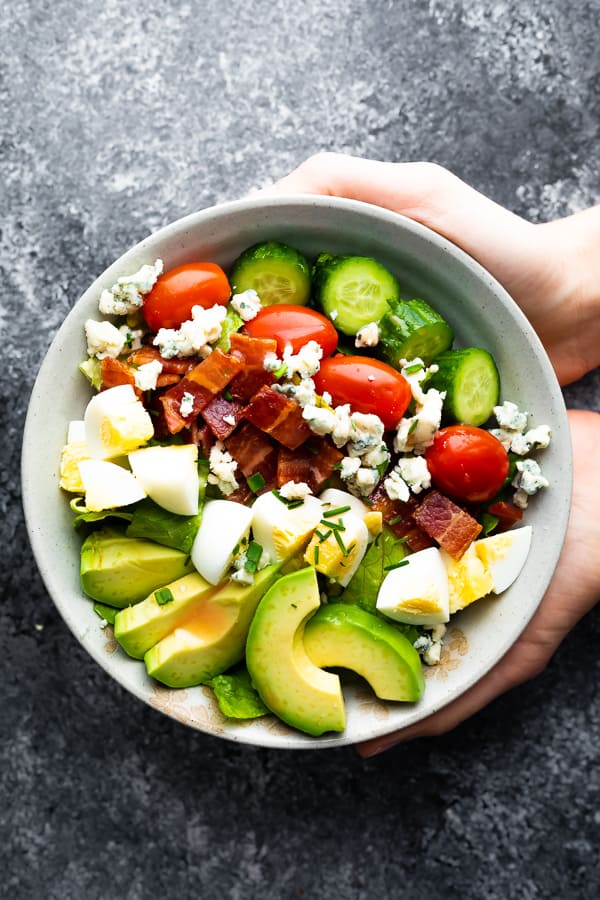 hands holding a gray bowl filled with cobb salad