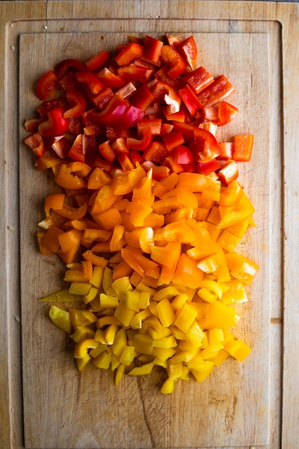 overhead view of red, orange and yellow bell peppers chopped into pieces on a wood cutting board
