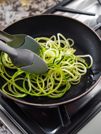 zucchini noodles being cooked in black pan on stove with tongs