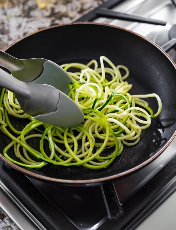 zucchini noodles being cooked in black pan on stove with tongs