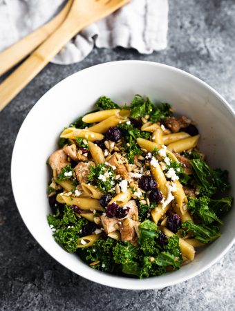 A large white bowl filled with kale chicken pasta salad with wood tongs in the background