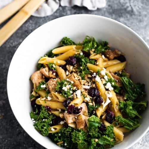 A large white bowl filled with kale chicken pasta salad with wood tongs in the background