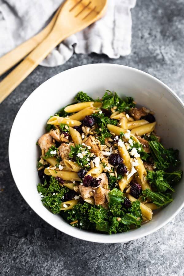 A large white bowl filled with kale chicken pasta salad with wood tongs in the background