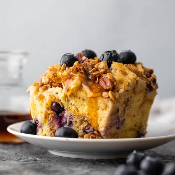 side view of a piece of french toast casserole on a white plate with blueberries on top and syrup behind it