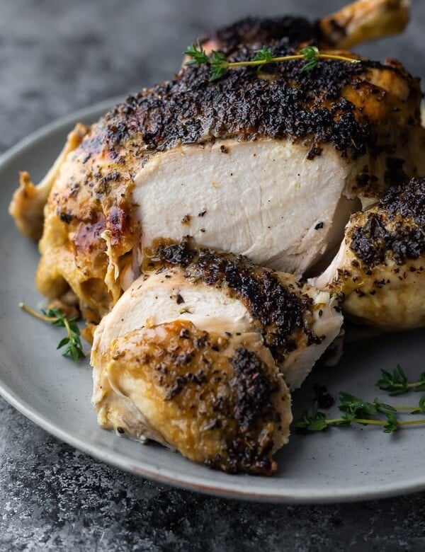 Close up shot of a whole cooked chicken with a few slices off on a gray plate with fresh thyme