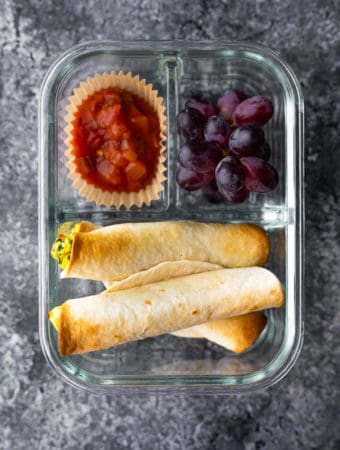 overhead shot of breakfast taquitos in a meal prep container with a side of salsa and grapes