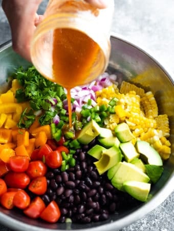 dressing being poured out of a mason jar over black bean and corn salad in large metal bowl