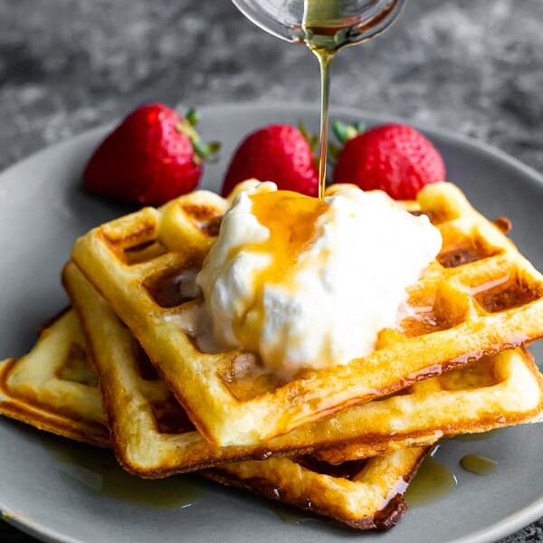 stack of three buttermilk waffles on a gray plate with syrup being drizzled over them and fresh strawberries